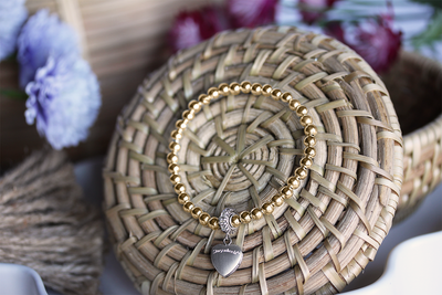 14k gold-filled stretch bracelet with sterling silver heart locket on basket surrounded by flowers. 