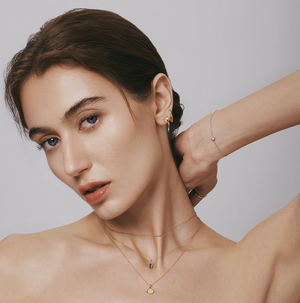 Woman wearing Aurelie Gi fine jewelry featuring 14k gold layered necklaces with diamond and sapphire pendants, dainty gold bracelet, and gemstone-accented hoop earrings, styled against a neutral background for an elegant minimalist look.