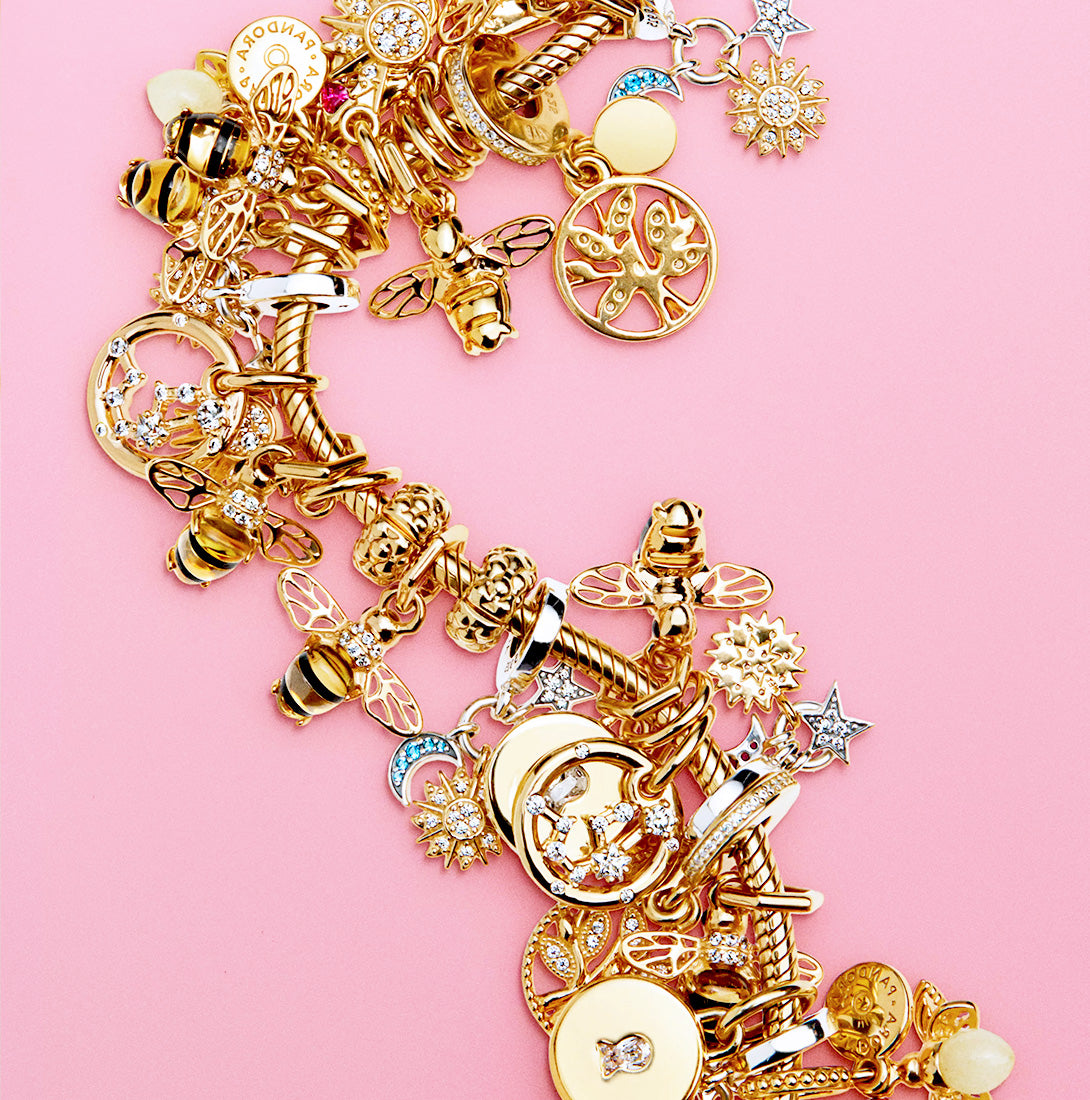 14k gold-filled Pandora Moments Smooth Snake Chain Bracelet adorned with exquisite charms including bees, zodiac symbols, a moon and stars motif, a key lock, and more, all set against a vibrant poppy pink background.