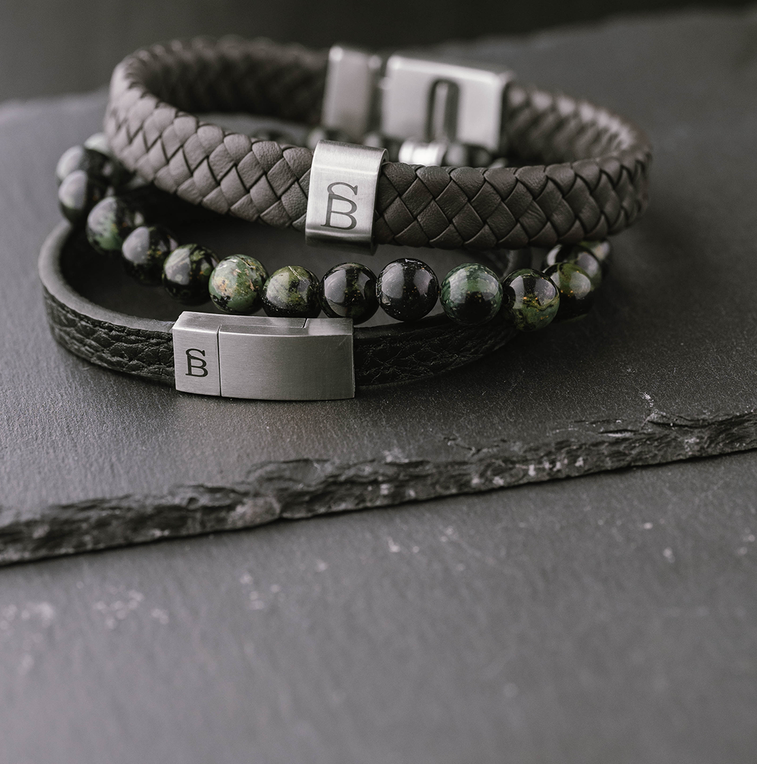 Close-up of men's stackable bracelets featuring black braided leather, green and black jasper beads, and stainless steel magnetic clasps engraved with a stylized 