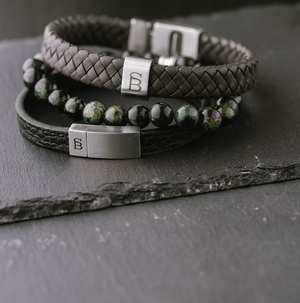 Close-up of men's stackable bracelets featuring black braided leather, green and black jasper beads, and stainless steel magnetic clasps engraved with a stylized "SB" logo, displayed on a dark slate surface.