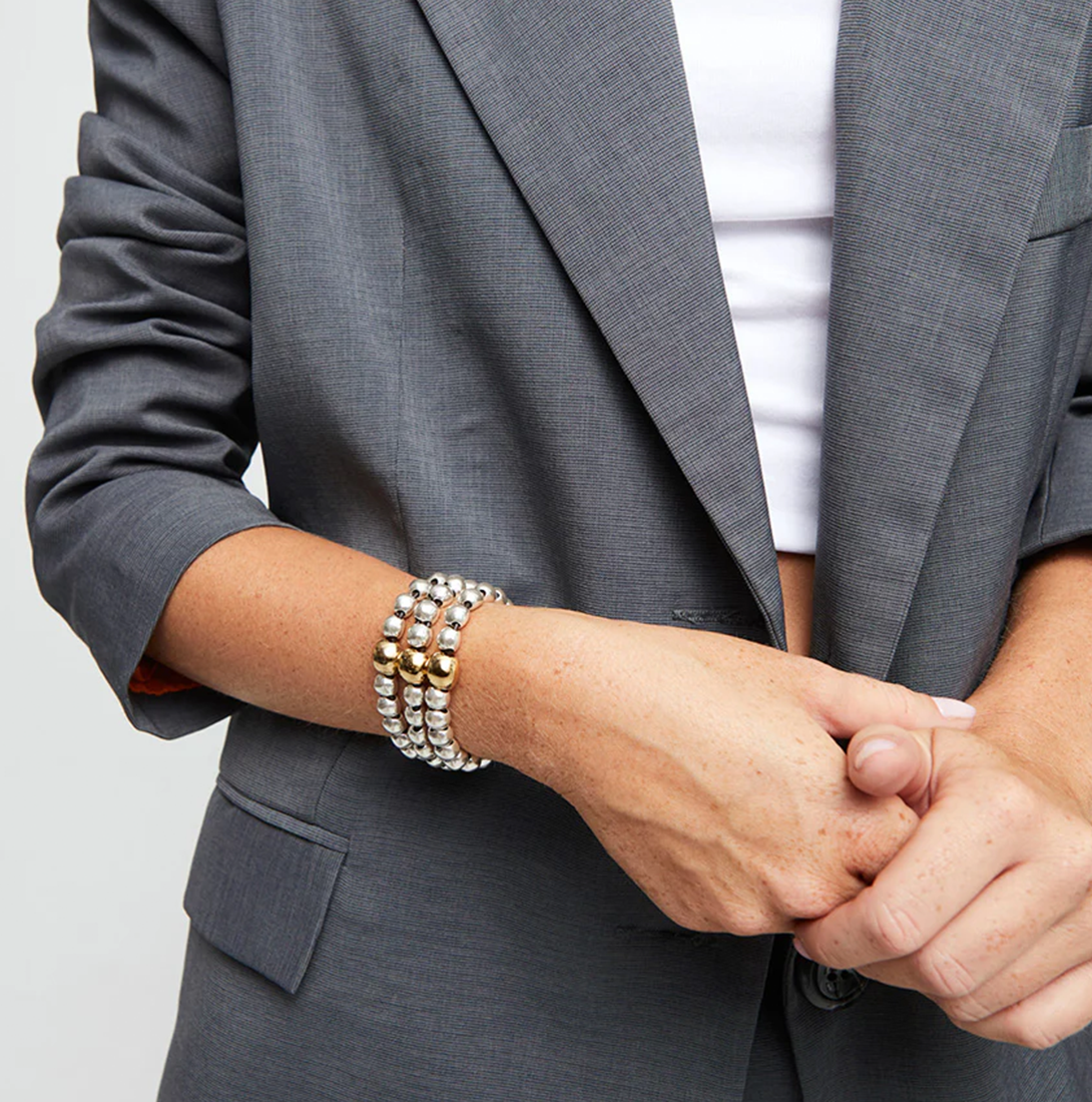 Stylish woman wearing a grey blazer accessorized with a bold multi-strand beaded bracelet featuring silver and gold-toned spheres by UNOde50.