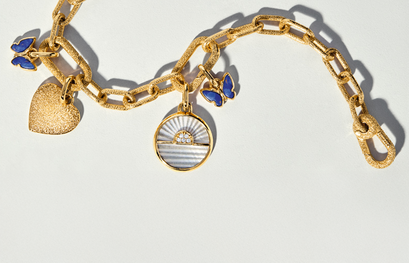 Pandora ME collection gold textured chain bracelet featuring customizable charms, including a glitter heart, enamel blue butterflies, and a circular sunset charm with pavé crystals, displayed on a light background