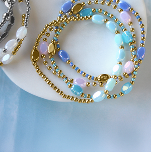 Layered stretchy waterproof bracelets featuring gold accents with stunning blue or pink beads, elegantly arranged on a white dish. Vibrant silver options with white beads complement the display, set against a soothing waterwash blue background.