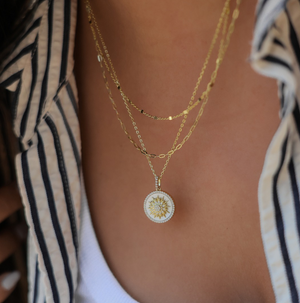 Lafonn jewelry layered gold chain necklaces featuring a round white enamel pendant with a gold sunburst and sparkling center stone, styled over a white tank top and striped shirt for a chic, modern look.