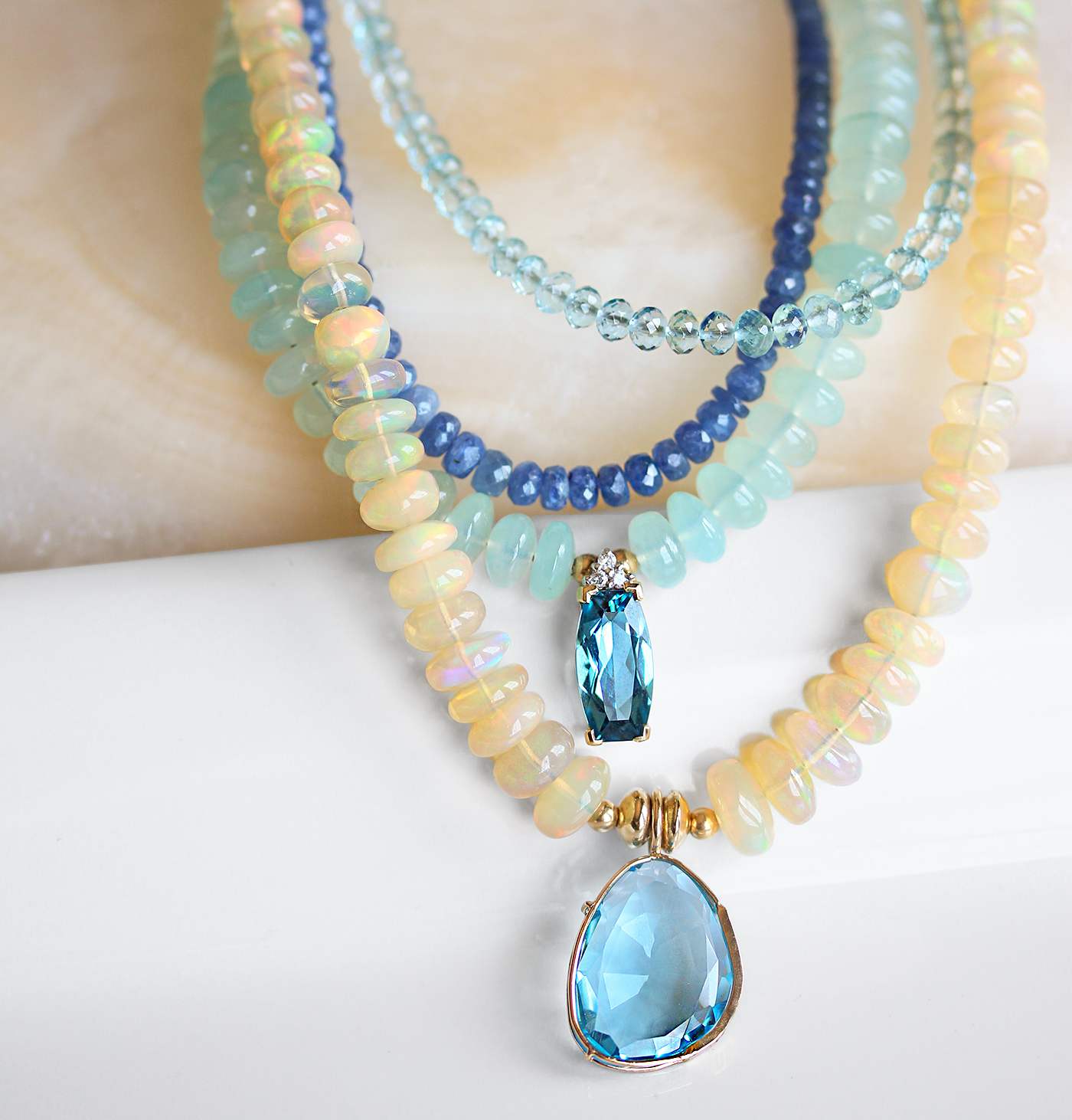 Close-up of layered gemstone necklaces featuring Ethiopian opals, aquamarine, kyanite, and blue topaz pendants — vibrant handcrafted jewelry with iridescent and faceted blue gemstones for a luxe coastal-inspired look.