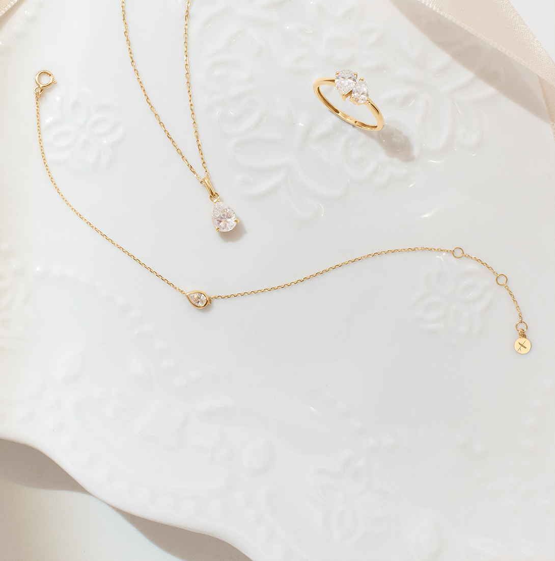 Aurelie Gi fine jewelry set in yellow gold featuring a pear-shaped solitaire necklace, matching delicate bracelet, and a three-stone pear and round-cut ring, displayed on an elegant white embossed ceramic plate.