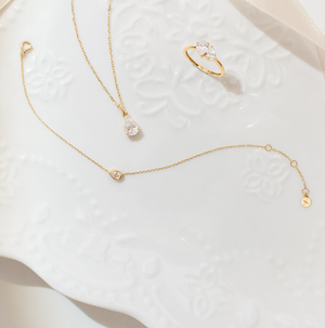 Aurelie Gi fine jewelry set in yellow gold featuring a pear-shaped solitaire necklace, matching delicate bracelet, and a three-stone pear and round-cut ring, displayed on an elegant white embossed ceramic plate.