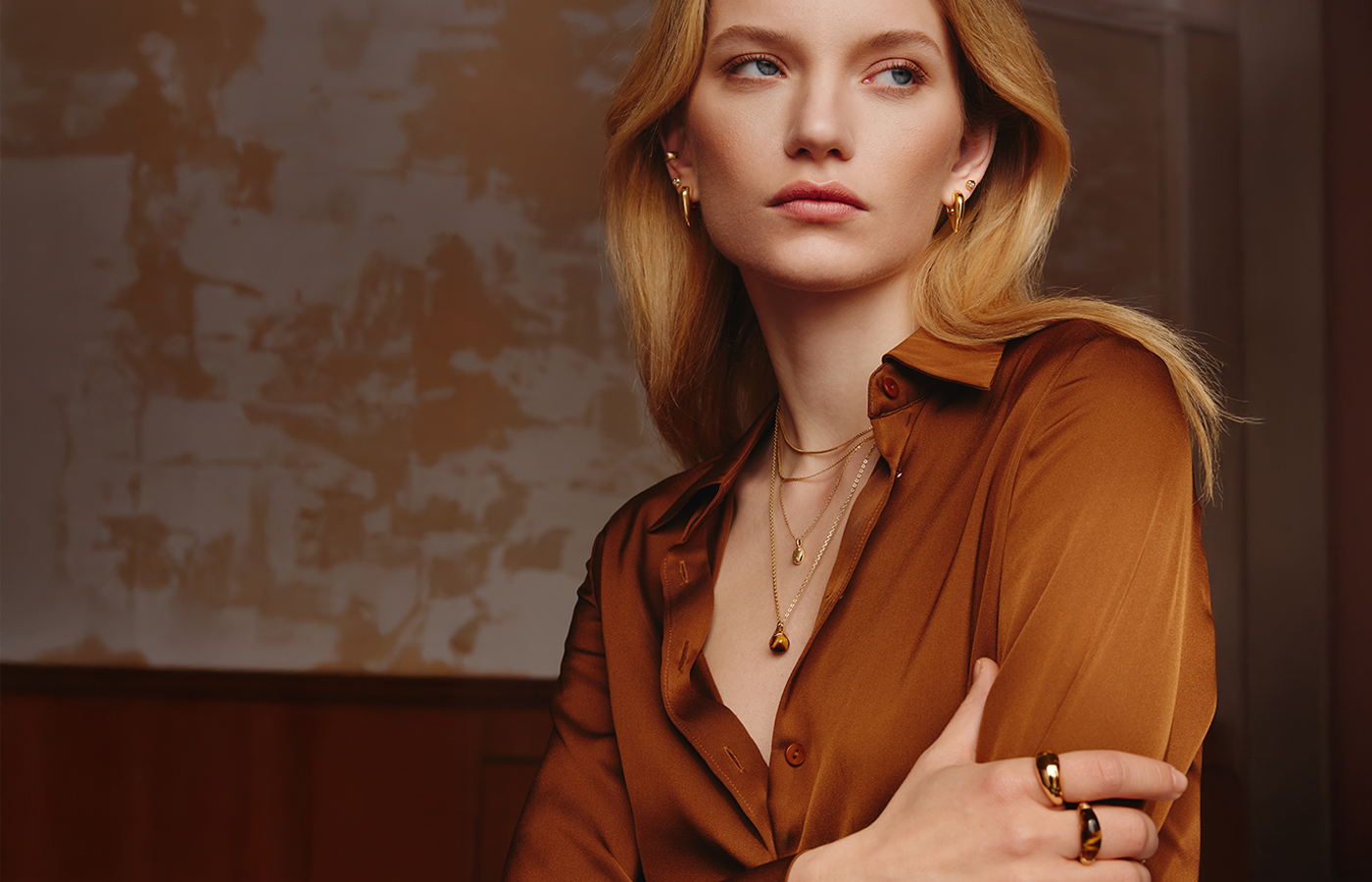 Model wearing Ania Haie Cozy Lady collection jewelry, including layered gold necklaces, statement gold rings, and chunky gold hoop earrings, styled with a warm brown satin blouse — modern autumn jewelry look from AW25 campaign.