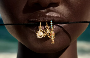 Pandora ocean-inspired gold charms featuring a pearl-studded shell, a starfish with crystal detail, and a colorful gemstone seahorse, displayed on a black cord held by a model’s lips with a beach background.