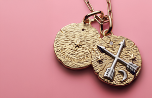 Pandora Talisman gold medallion necklace featuring crossed arrows, stars, and crescent moon design on textured gold discs, displayed against a soft pink background.
