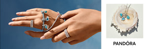 Pandora ocean-themed jewelry collection featuring silver charm bracelets with blue crystal dolphin and wave charms, pavé band rings, and a shell displaying matching sea-inspired pendant necklaces with turquoise and aqua accents, set against a coastal blue sky and seashell backdrop.