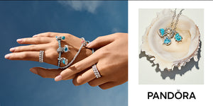 Pandora ocean-themed jewelry collection featuring silver charm bracelets with blue crystal dolphin and wave charms, pavé band rings, and a shell displaying matching sea-inspired pendant necklaces with turquoise and aqua accents, set against a coastal blue sky and seashell backdrop.