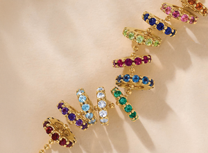 Gold birthstone charms featuring vibrant round-cut gemstones in every color of the rainbow, symbolizing each month, displayed on a neutral background.