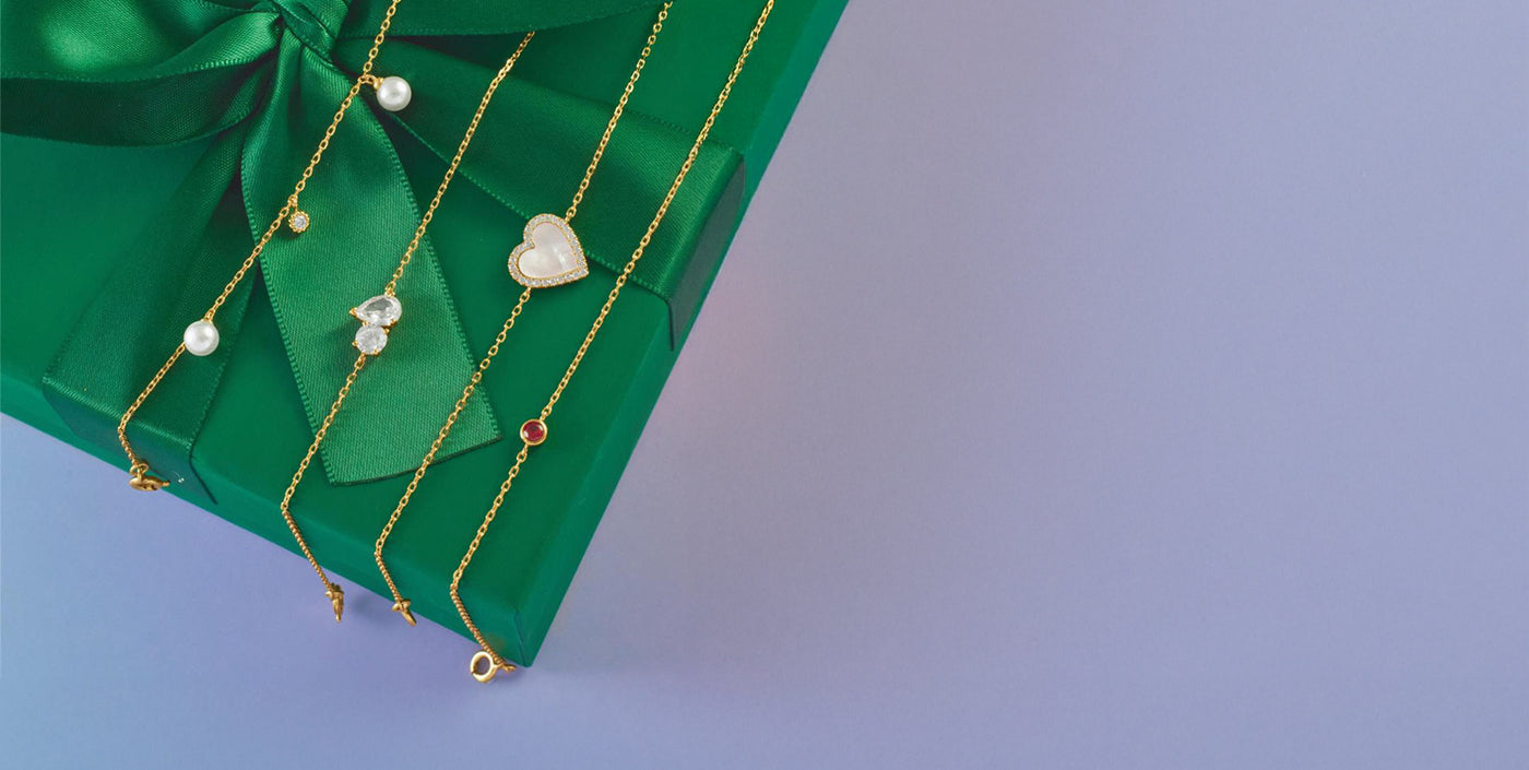 Winter jewelry gift inspiration featuring delicate gold chain necklaces with pearls, hearts, and gemstones displayed on a green ribbon-wrapped box — shop elegant new arrivals for the season.