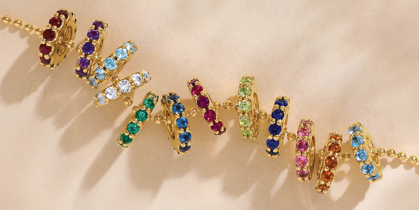 Gold birthstone charms featuring vibrant round-cut gemstones in every color of the rainbow, symbolizing each month, displayed on a neutral background.